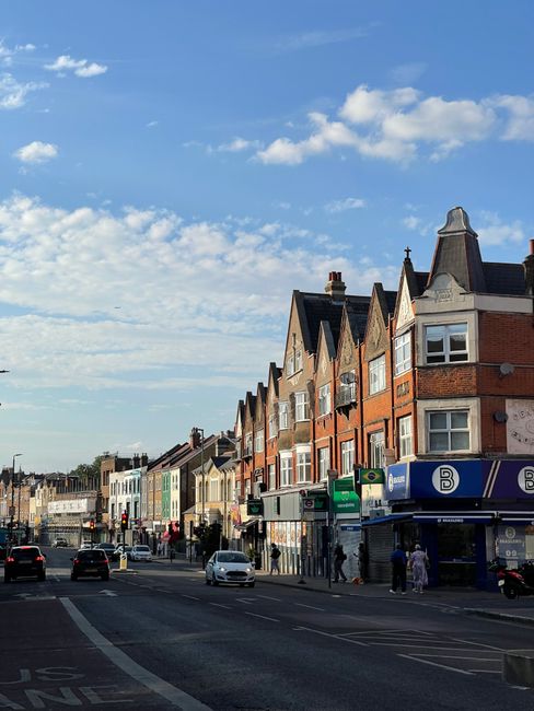 A street scene in Tufnell Park showing a row of multi-story terraced houses with brick facades, some featuring bay windows and sloped roofs. The pavement is occupied by pedestrians walking past the shops and residential entrances, with a few people awaiting or boarding buses at a nearby bus stop. A commercial vehicle, likely used for moving or furniture transport, is partially visible parked on the street, near the curb. Several cars are driving along the road, which has marked lanes and traffic lights. The sky above is partly cloudy with patches of blue, indicating daytime. The environment suggests a vibrant urban area suitable for home relocation and moving services, as provided by Tufnell Park Man and Van, with nearby shops and residential buildings making it a typical setting for household packing and furniture transport activities.
