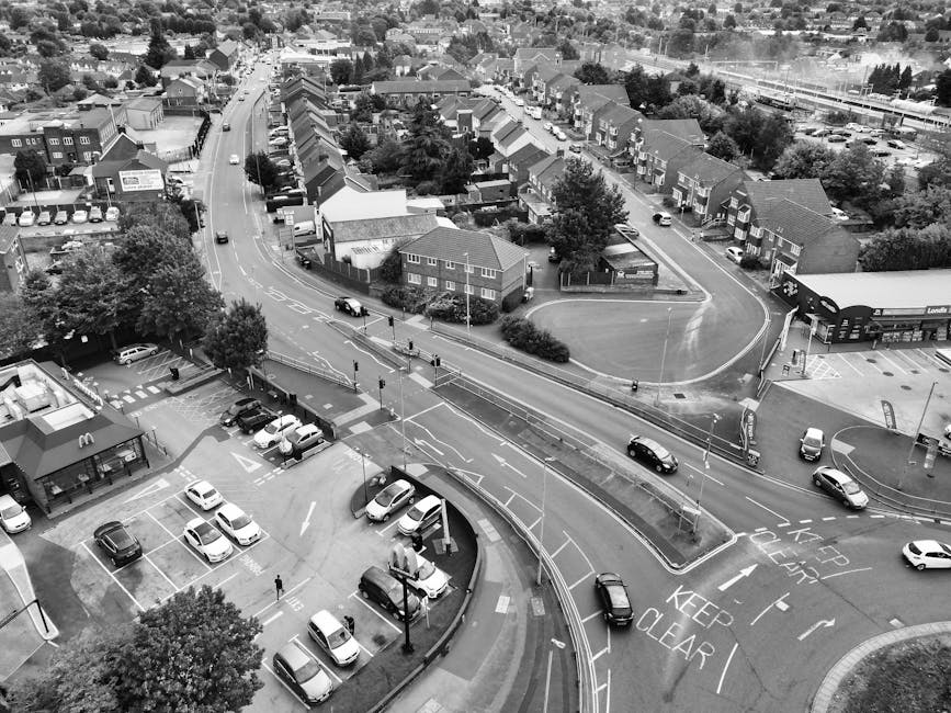 A black and white aerial photograph showing a street scene with a parking lot in the foreground containing several parked cars, some open car doors, and a McDonald's restaurant with outdoor seating area visible. A bus stop with a shelter and a few pedestrians walking nearby are also present. In the middle ground, a curved road with moving vehicles such as cars and a van is marked with road signs including 'Keep Clear' and lane markings. The background features a residential area with rows of terraced houses, some with small front gardens, and trees lining the streets. There is no visible furniture, packing materials, or storage items; the scene captures urban infrastructure and vehicle traffic, relevant to the logistics involved in house removals or local moving services, especially around areas like Junction Road in Tufnell Park as provided by Tufnell Park Man and Van.