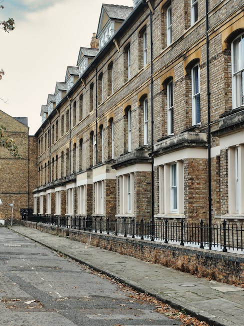A row of three-storey Victorian brick terraced houses with arched windows and white window frames, situated along a paved street in Tufnell Park. In front of the houses, there is a narrow sidewalk with a low brick wall topped by a black metal railing. The houses have small front gardens with some foliage, and the street appears quiet with no vehicles or pedestrians present. The scene is captured during daylight hours with overcast skies, and the building's architectural details are clearly visible. This setting reflects a typical residential area suitable for house removals or property relocations, with the street ready for loading and unloading of furniture and boxes by Tufnell Park Man and Van during a home relocation process.