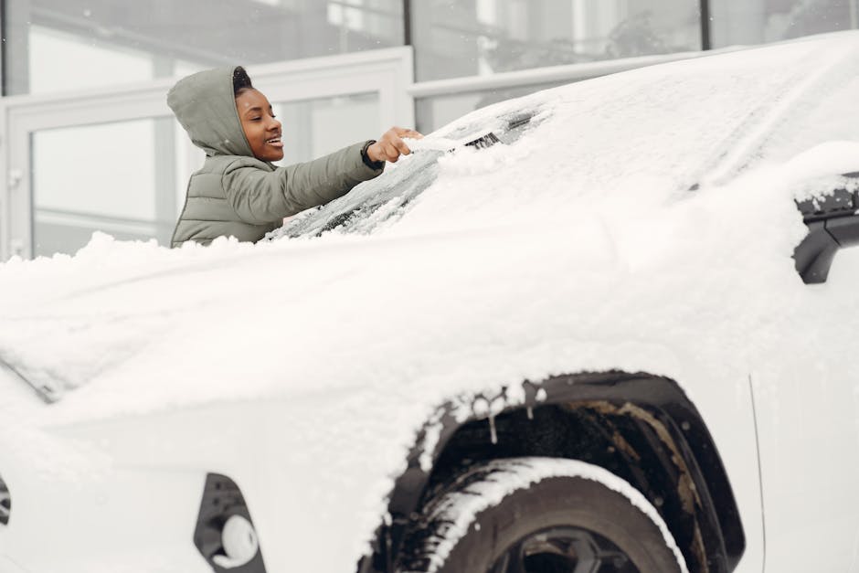 A person wearing a grey hooded jacket is clearing snow from the roof of a white vehicle, likely a van or small moving truck, parked outdoors in front of a modern building with large glass windows. The individual is using their hands to brush off accumulated snow, focusing on the curved surface of the vehicle's roof. The vehicle's wheel is visible, partially covered in snow, and the environment appears cold, possibly during a home relocation or moving process affected by winter conditions. This scene illustrates the importance of preparing moving equipment and vehicles, such as vans, for winter weather during furniture transport or home relocation tasks, and is associated with professional removals services from Tufnell Park Man and Van.